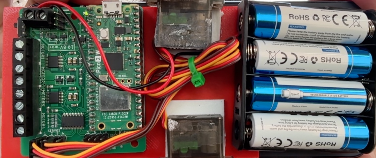 Top-down layout on the plate: carrier board with the Pico on one side, both servos flat in the middle with shafts out, the 4xAA battery pack on the other side.