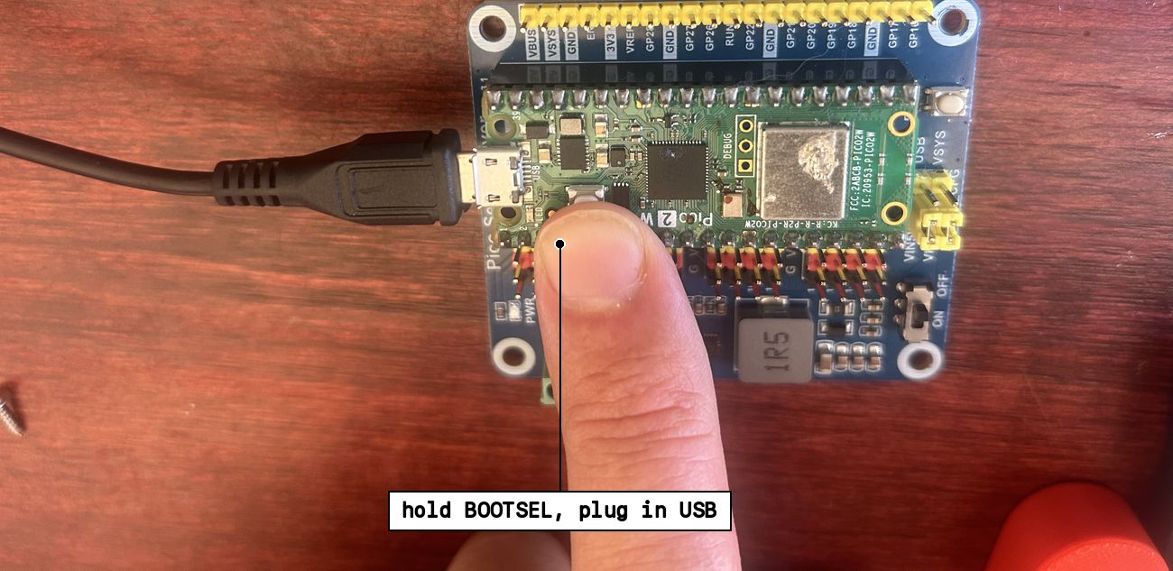 A finger holding the BOOTSEL button down while a USB cable is plugged into the Pico's port, the board still seated on the carrier.